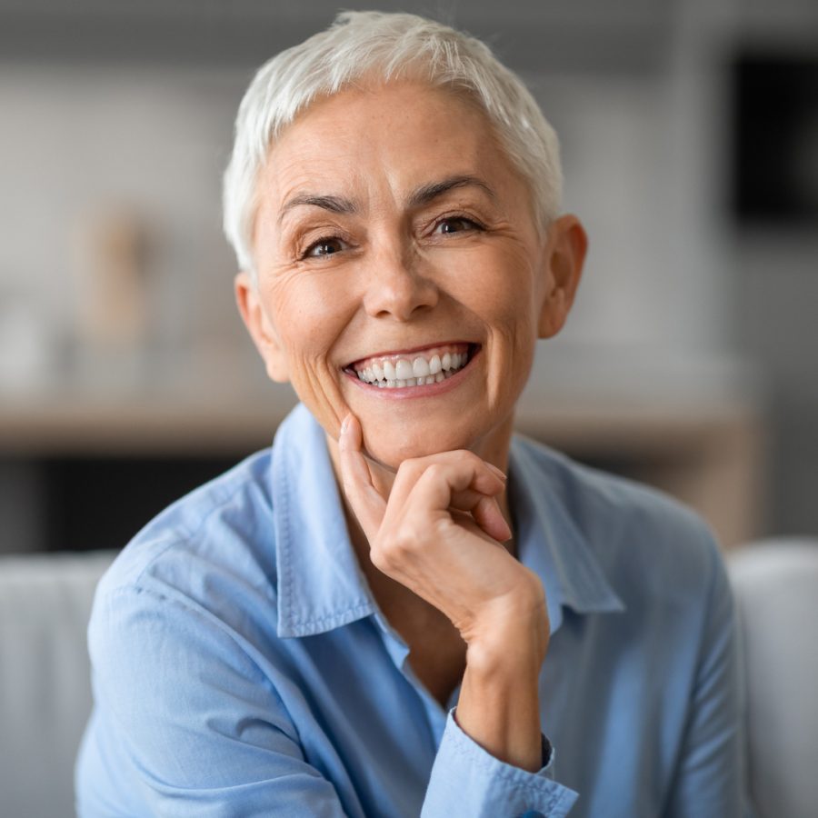 Portrait of lovely older gray woman smiling to camera and touching chin, sitting on sofa at home indoors. Happy beautiful mature lady resting and relaxing in living room, expressing happiness